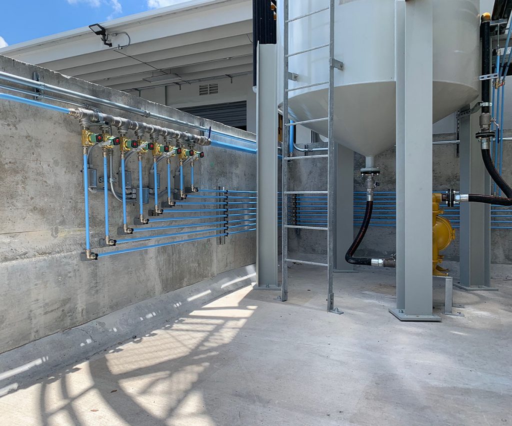 Industrial setting showcasing neatly organized piping and conduit runs against a concrete wall, with cylindrical tanks and structural supports from Arle Compressors of Florida under a clear blue sky.