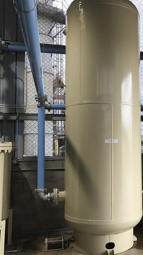 Industrial setting featuring a tall beige cylindrical tank with piping connected at the top and bottom, situated near wire mesh safety barriers, belonging to Arle Compressors of Florida.