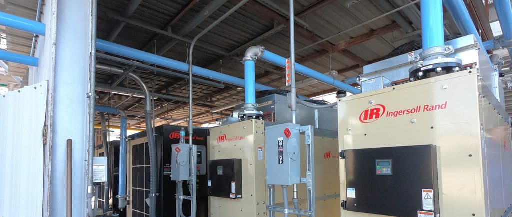 An industrial setting showcasing a row of large Arle Compressors of Florida air compressors and related machinery installed inside a facility with an array of pipes overhead.